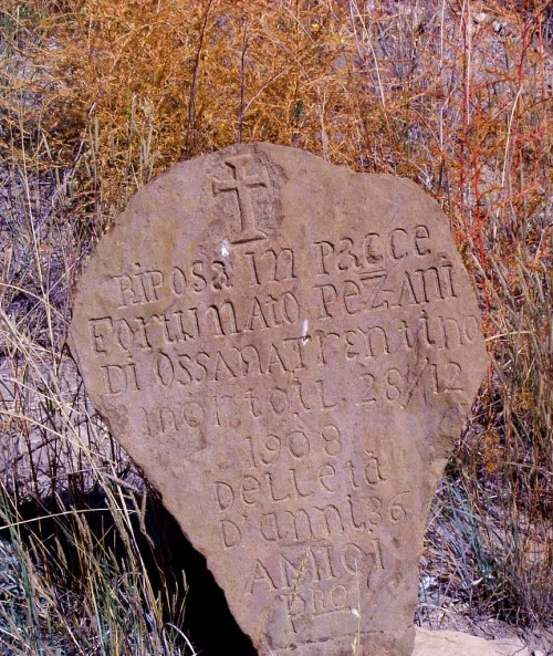 A weathered stone grave marker with an inscription in Italian, featuring a cross and surrounded by dry grass.