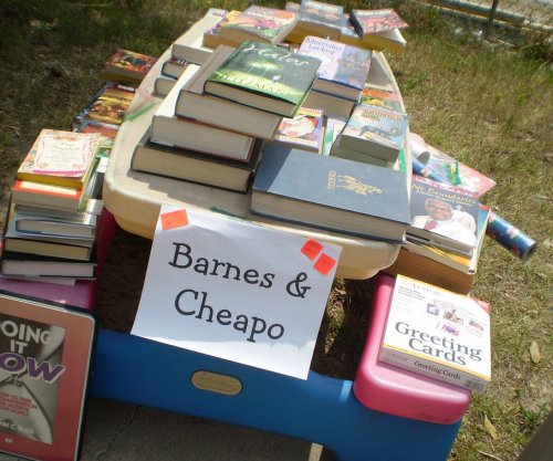 A table outdoors displaying a variety of books and greeting cards, with a sign reading "Barnes & Cheapo."