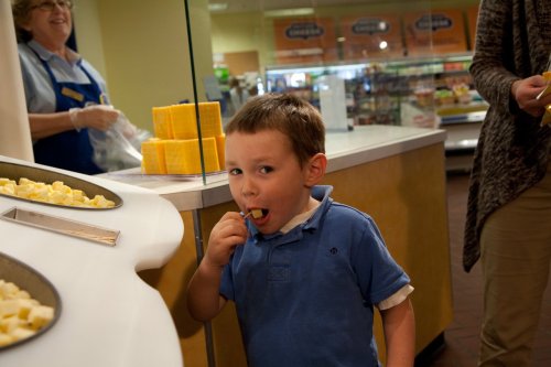 A young boy tastes cheese samples at a grocery store, smiling while a staff member prepares more samples in the background.