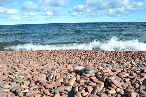 A rocky beach with smooth stones, gentle waves lapping at the shore, and a blue sky dotted with clouds.