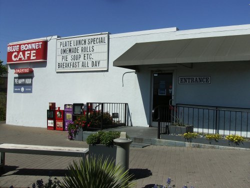 Exterior of Blue Bonnet Cafe featuring a sign with menu specials and an entrance with a ramp and flower planters.