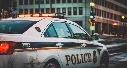 A police car with flashing lights parked on a city street, with buildings and traffic signals in the background.