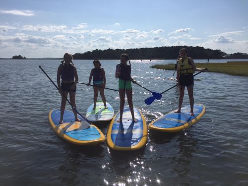 Four girls stand on paddleboards in a calm waterway, enjoying a sunny day with blue skies and greenery in the background.