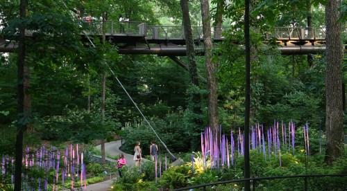 A lush green park with a walkway above and colorful flowers below, featuring people walking along the paths.