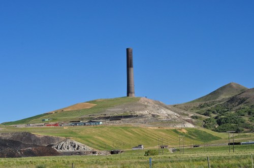 A tall smokestack rises from a green hillside, surrounded by rolling terrain and a clear blue sky.