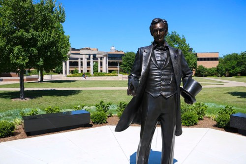 A bronze statue of Abraham Lincoln stands in a green park with trees and buildings in the background under a clear blue sky.