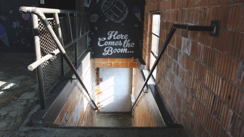A staircase leading down, illuminated by sunlight, with a chalkboard wall that reads "Here Comes the Boom..."