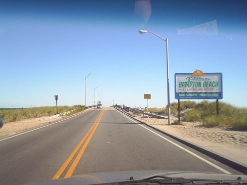 A road leading to Hampton Beach, with a welcome sign and sandy surroundings under a clear blue sky.