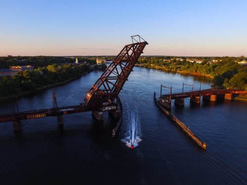 Aerial view of a raised railway bridge over a river, with a small boat passing underneath in clear blue skies.