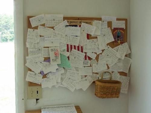 A bulletin board cluttered with various notes, cards, and messages, with a small basket attached.