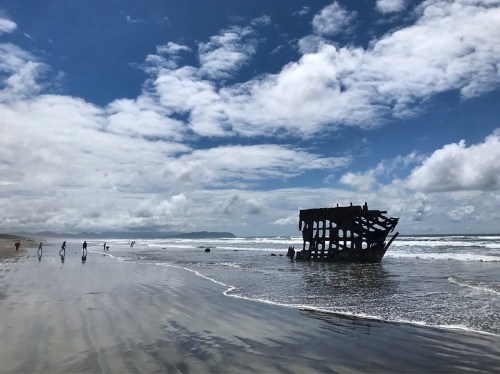 A shipwreck on a sandy beach under a cloudy sky, with people walking along the shore and waves crashing nearby.