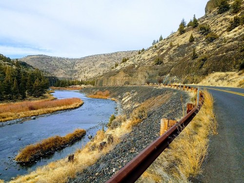 A winding road alongside a river, surrounded by hills and sparse trees, under a cloudy sky.