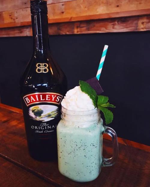 A mint green milkshake topped with whipped cream and a mint leaf, next to a bottle of Baileys Irish Cream.