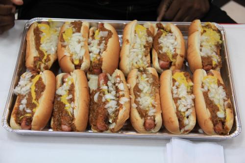 A tray of hot dogs topped with chili, mustard, and chopped onions, arranged neatly in rows.
