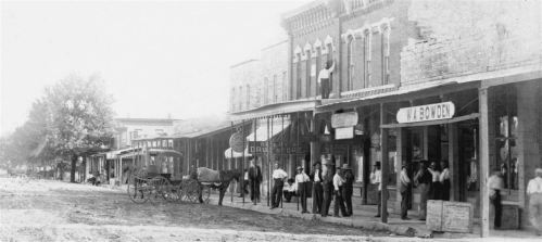Black and white photo of a historic street scene with storefronts, a horse-drawn carriage, and people gathered outside.