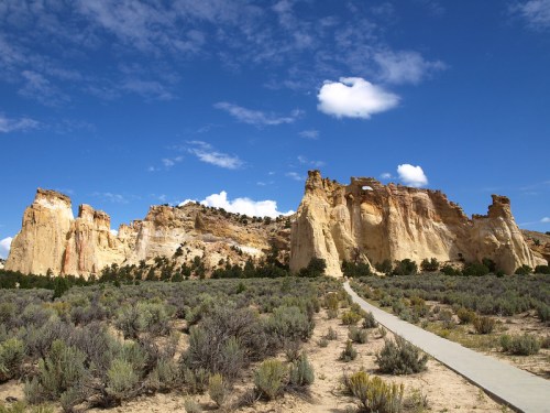A scenic landscape featuring towering rock formations under a blue sky with scattered clouds, surrounded by sagebrush.