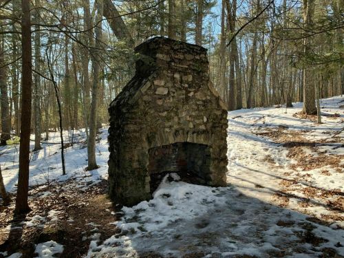 A stone chimney stands in a snowy forest, surrounded by trees and dappled sunlight.