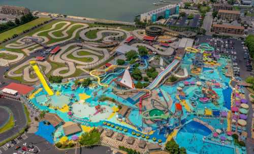 Aerial view of a vibrant water park with slides, pools, and go-kart tracks near a lake, bustling with visitors.