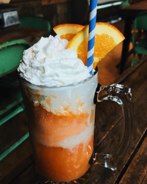 A frosty orange drink topped with whipped cream and orange slices, served in a glass mug with a blue striped straw.
