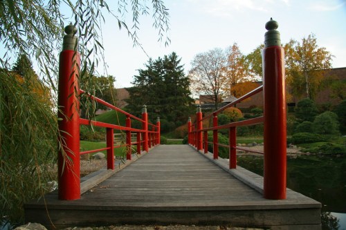A wooden bridge with red railings spans a tranquil pond, surrounded by trees and autumn foliage.