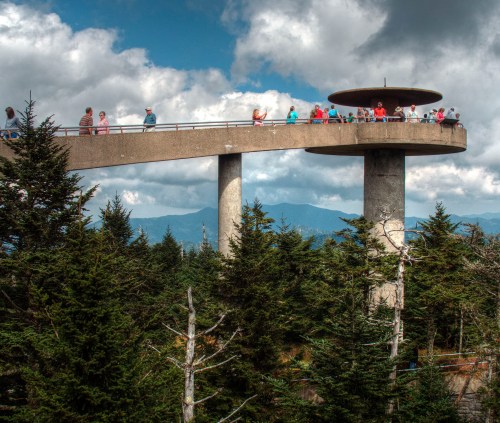 A concrete observation deck atop a forested area, with people enjoying the view against a backdrop of clouds and mountains.