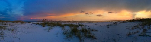 A panoramic view of a beach at sunset, featuring colorful skies and gentle waves, with grass in the foreground.