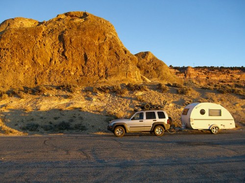 A silver SUV parked beside a small white camper trailer against a rocky landscape at sunset.