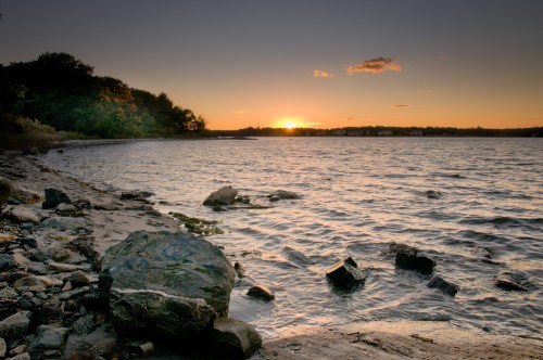 Sunset over a calm water body, with rocky shoreline and trees in the background, creating a serene landscape.