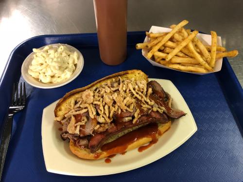 A plate with a loaded sandwich, fries, and macaroni salad on a blue tray. The sandwich is topped with meat and crispy onions.