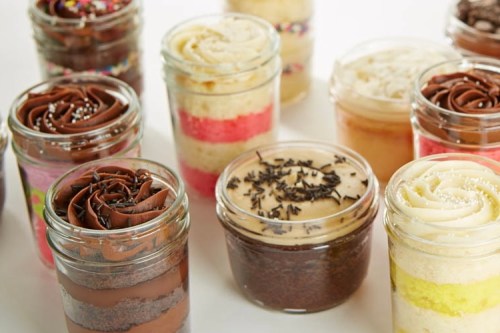 A variety of colorful layered cakes in jars, topped with swirls of frosting and sprinkles.