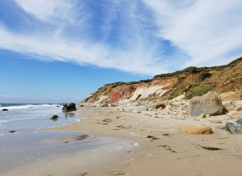 A sandy beach with large rocks and cliffs under a blue sky with wispy clouds. Waves gently lap at the shore.