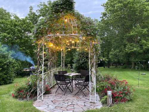 A charming garden gazebo adorned with lights and flowers, surrounded by lush greenery and a cozy dining setup.
