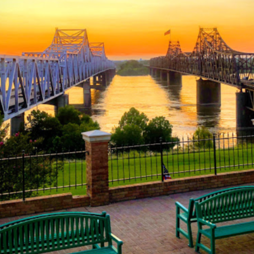 Sunset view of two bridges over a river, with benches in the foreground and lush greenery along the banks.
