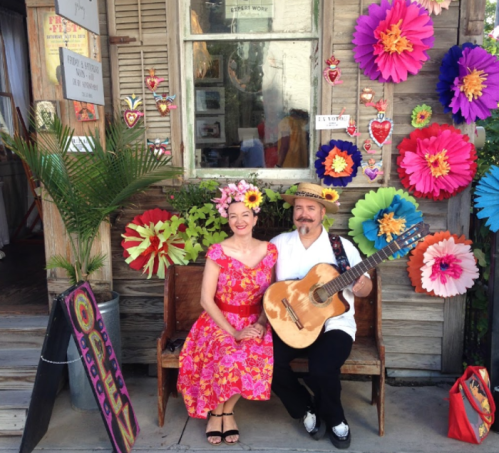 A woman in a colorful dress and a man with a guitar sit on a bench surrounded by vibrant paper flowers.