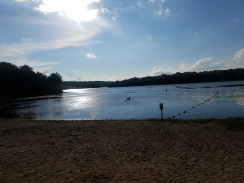 A serene lake scene with a sandy beach, swimmers in the water, and a bright sky with scattered clouds.