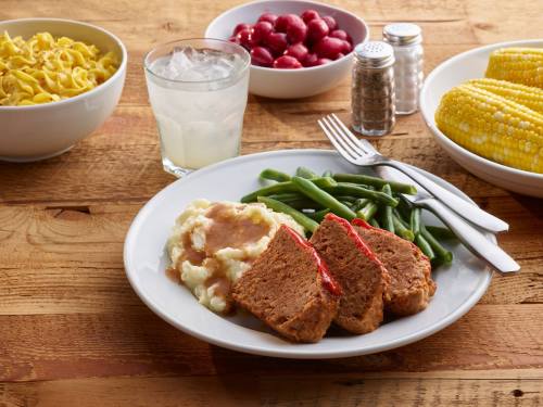 A plate with meatloaf, mashed potatoes, green beans, and corn, accompanied by bowls of noodles and cherries, and a glass of water.
