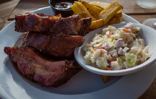 A plate of barbecue ribs with coleslaw, fried plantains, and a small cup of sauce on the side.