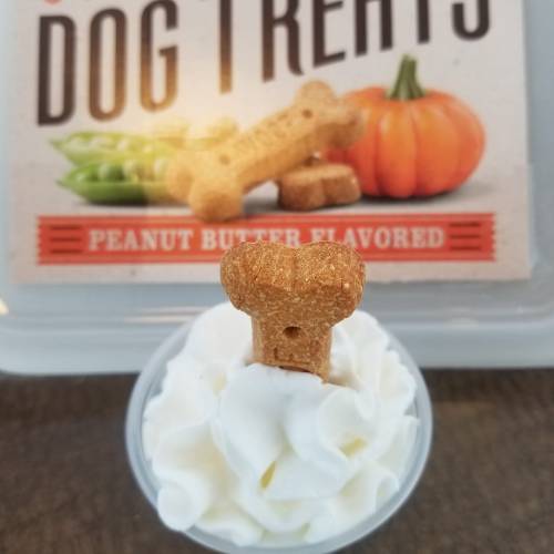 A peanut butter dog treat balanced on top of a swirl of whipped cream, with a package of dog treats in the background.
