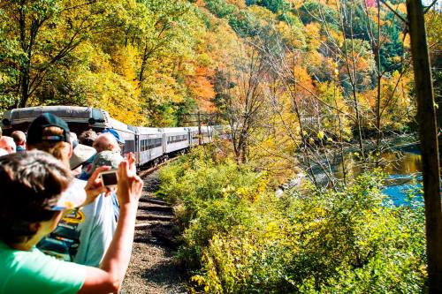 A train travels along a scenic route with colorful autumn foliage, while onlookers take photos from the side.