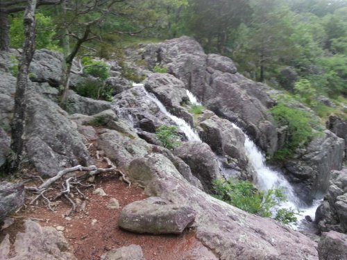 A rocky landscape with a small waterfall cascading down, surrounded by lush greenery and trees.