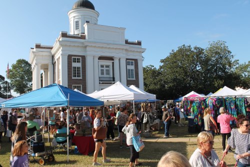 A bustling outdoor market with tents and vendors in front of a historic building, filled with people browsing and shopping.