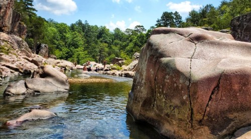 A serene river scene with large rocks, surrounded by lush greenery and people enjoying the outdoors under a blue sky.