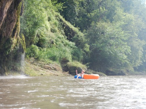 A person in an orange and blue inflatable tube floats on a river, surrounded by lush greenery and a waterfall.