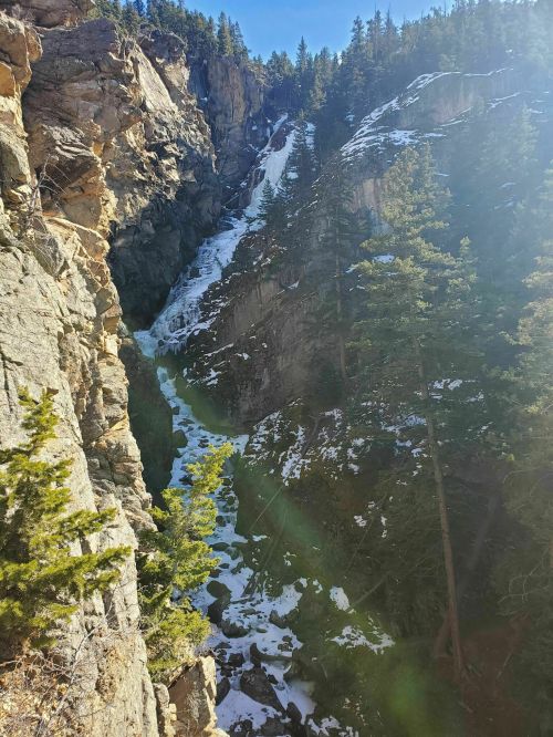 A scenic view of a rocky canyon with a river flowing through, surrounded by evergreen trees and snow-capped cliffs.