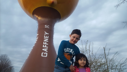 A boy and girl stand in front of a large water tower in Gaffney, SC, under a cloudy sky.