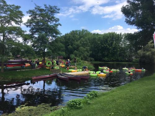 A lively scene by a river with people in colorful kayaks and inflatables, surrounded by greenery and blue skies.
