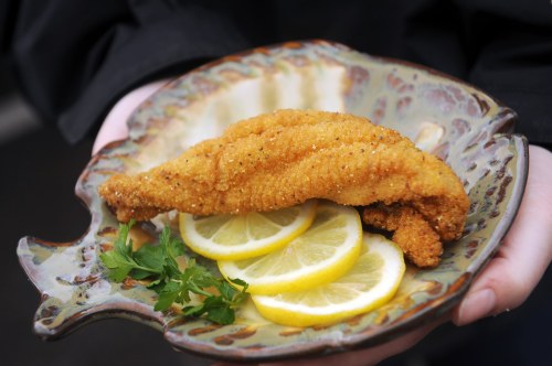 A person holds a decorative plate with a fried fish fillet and lemon slices, garnished with fresh parsley.