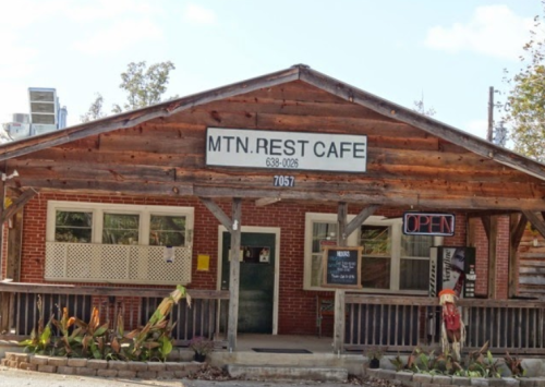 A rustic wooden café with a sign reading "MTN. REST CAFE" and an "OPEN" sign, surrounded by greenery.