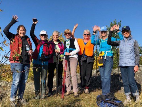A group of eight women hikers, smiling and waving, standing outdoors against a clear blue sky.