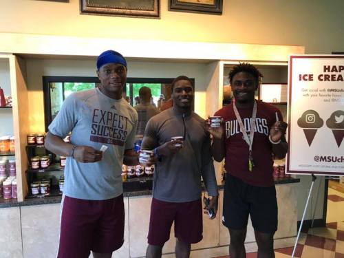 Three athletes in casual sportswear hold ice cream cups, smiling in a brightly lit room with a promotional sign.